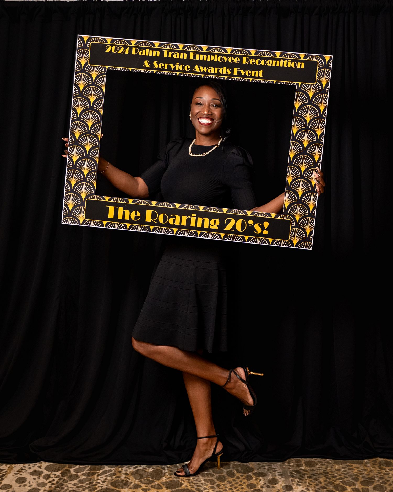 A guest poses in a black dress with a decorative art deco photo frame against a dark background.