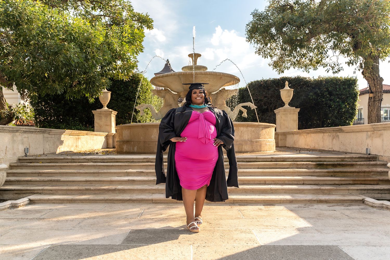 Graduate in academic robes and pink dress walks down stone steps near fountain.