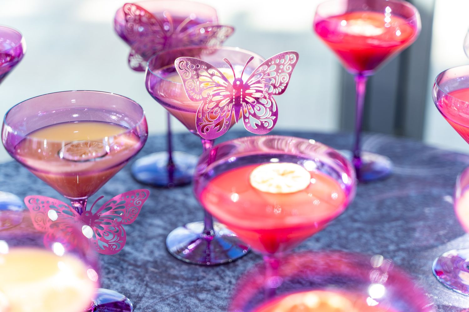 Pink cocktails garnished with decorative purple butterflies on dark surface with candlelight.