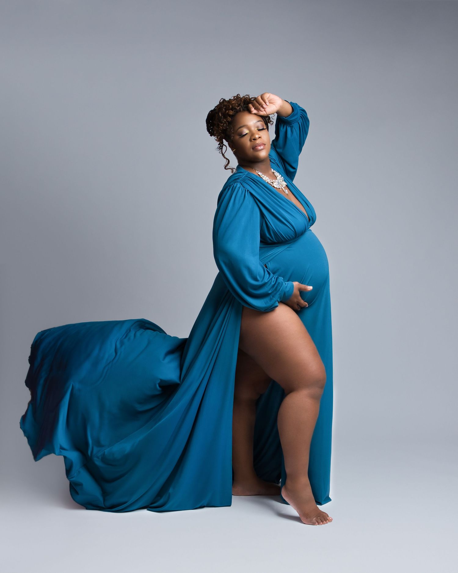 Elegant maternity photoshoot featuring a long blue dress with flowing fabric against light gray backdrop.