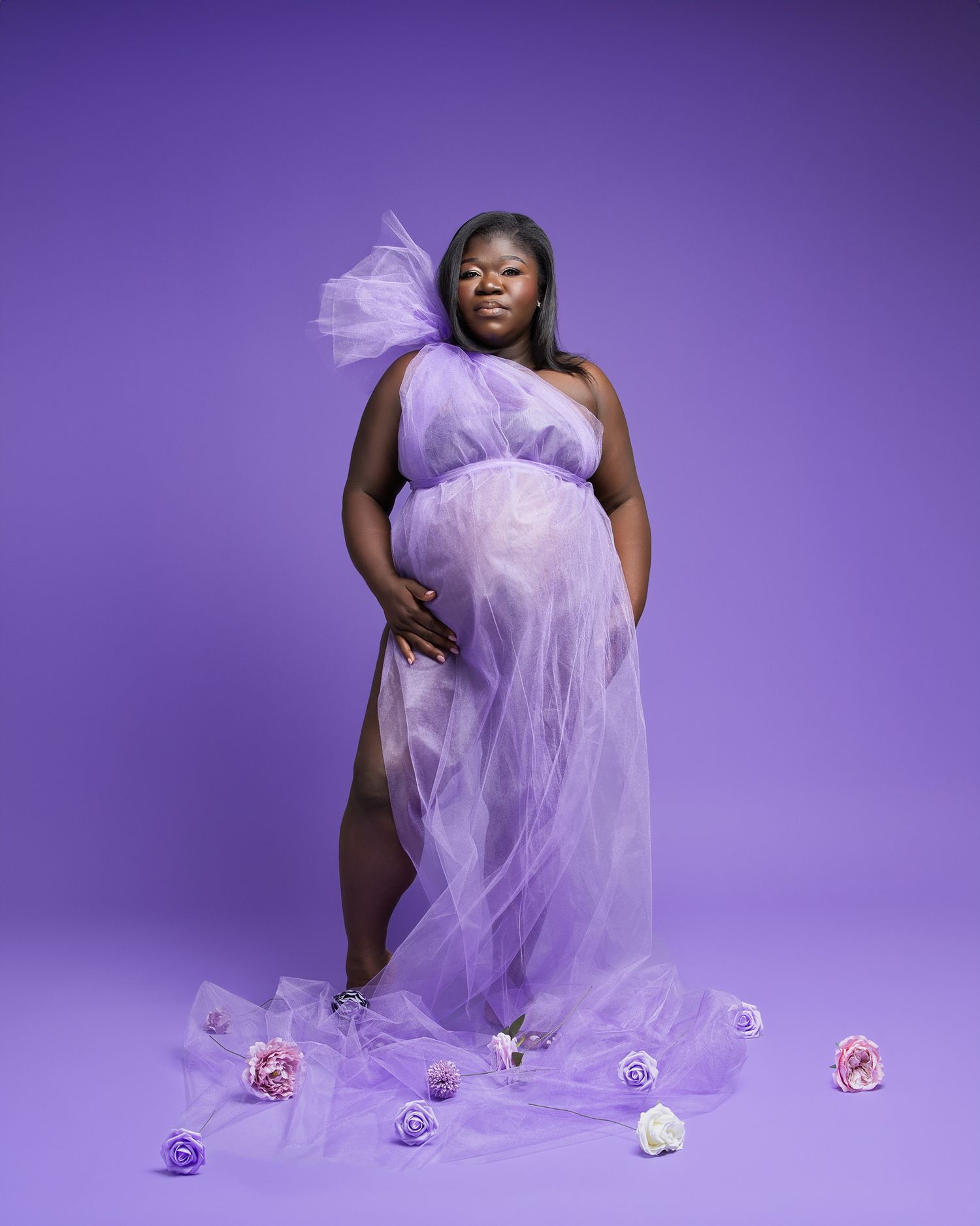 Maternity photo with flowing purple tulle dress and scattered flowers against purple backdrop.