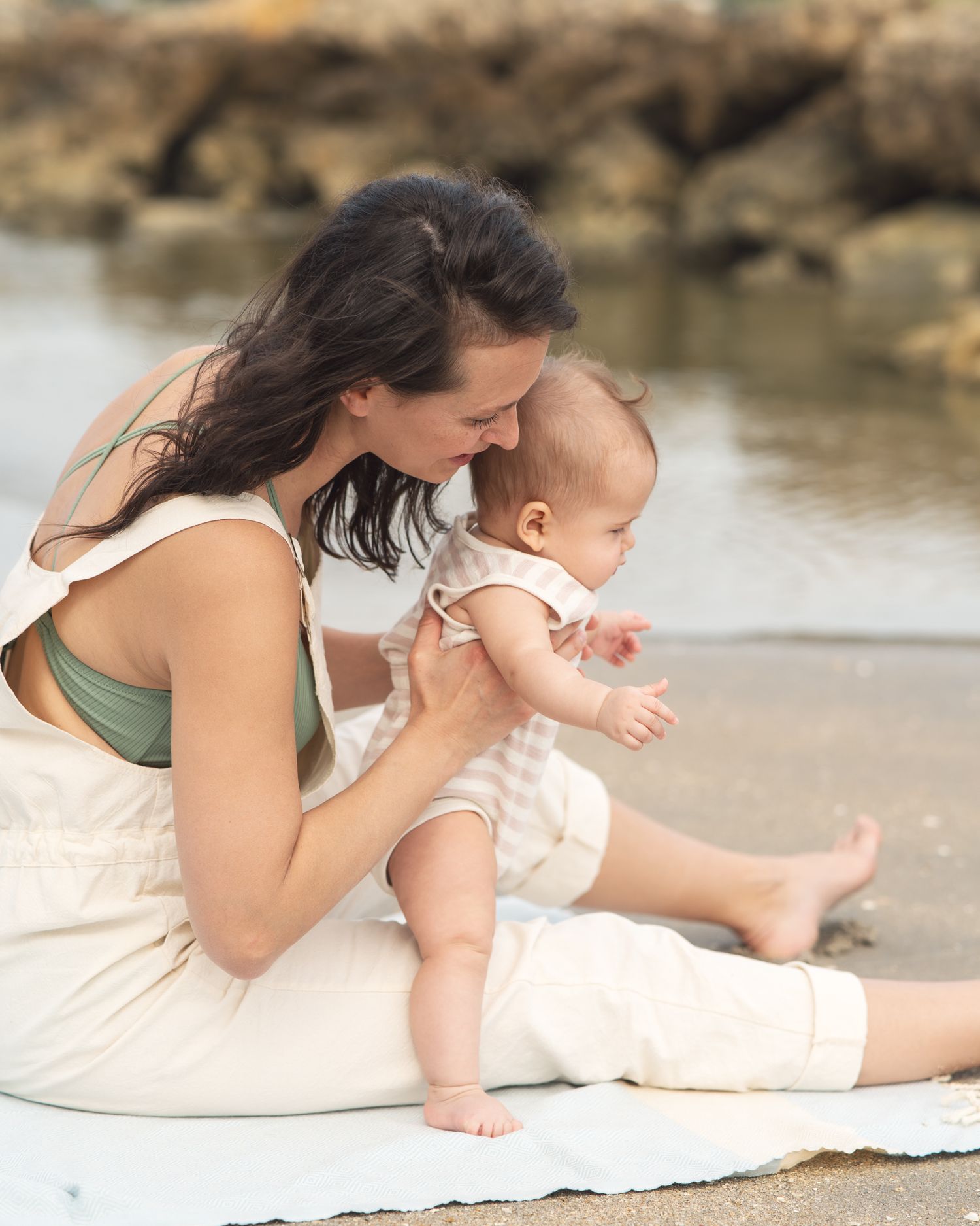A parent and baby share a quiet moment sitting on a blanket by the water's edge.