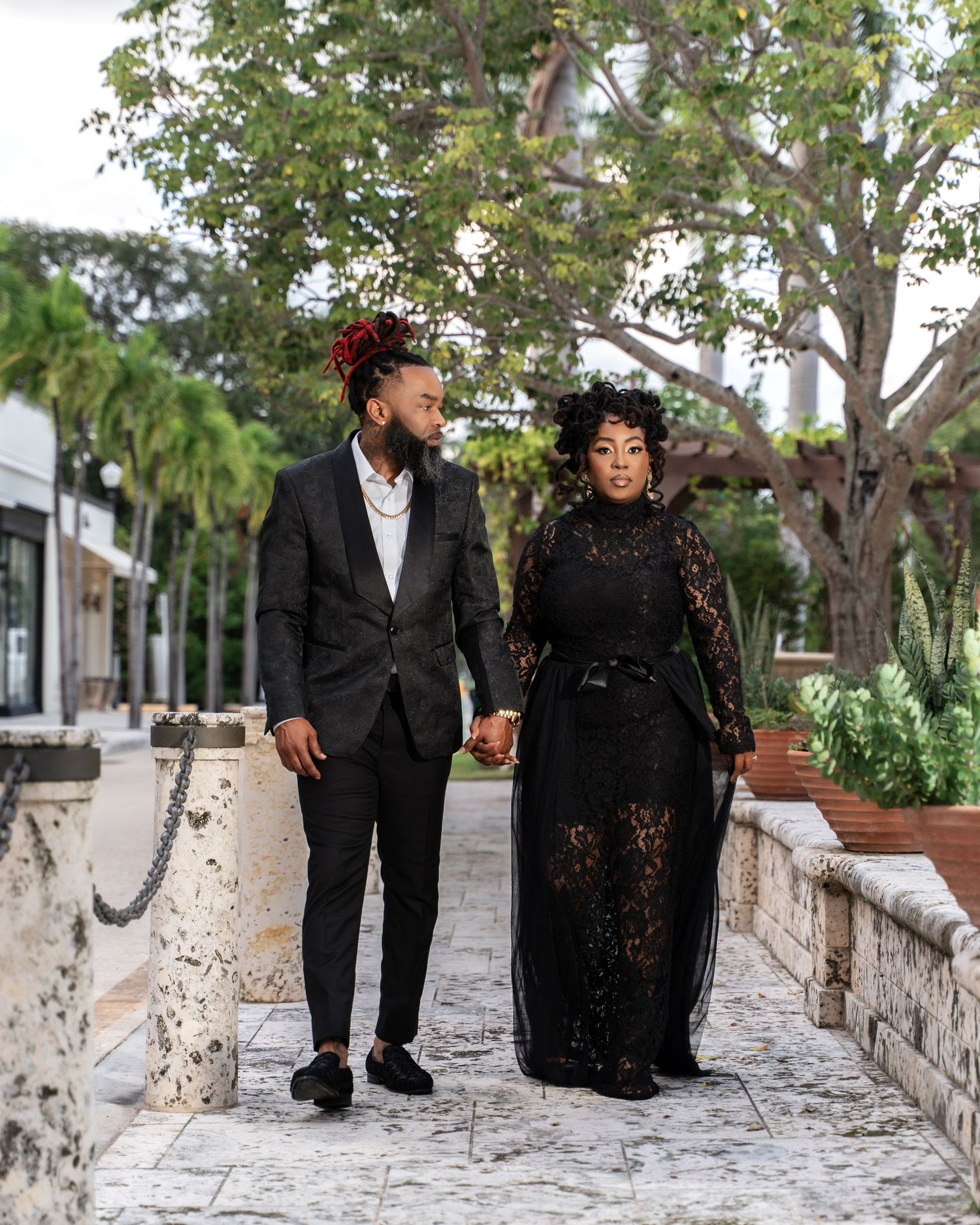 A couple in formal black attire walks together along a stone pathway surrounded by lush tropical landscaping.