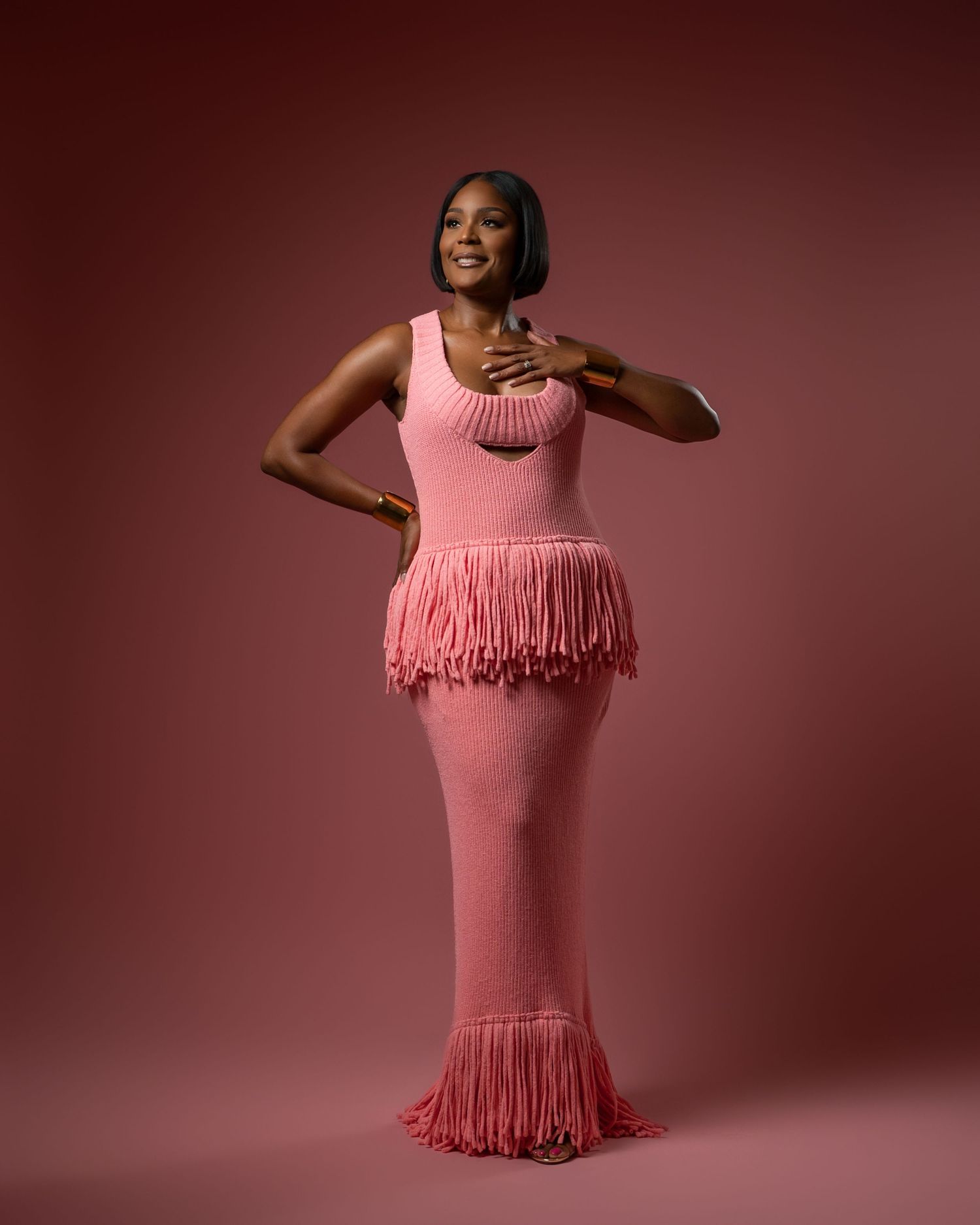 A model poses in a pink fitted dress with fringe details at the waist and hem against a burgundy background.