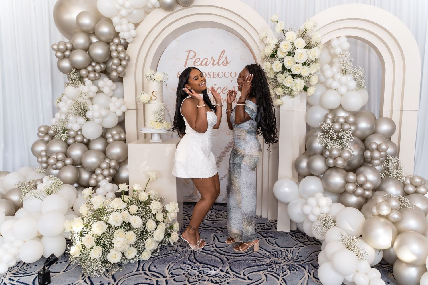Two women pose together in front of an elegant white balloon arch and floral display at a special event.