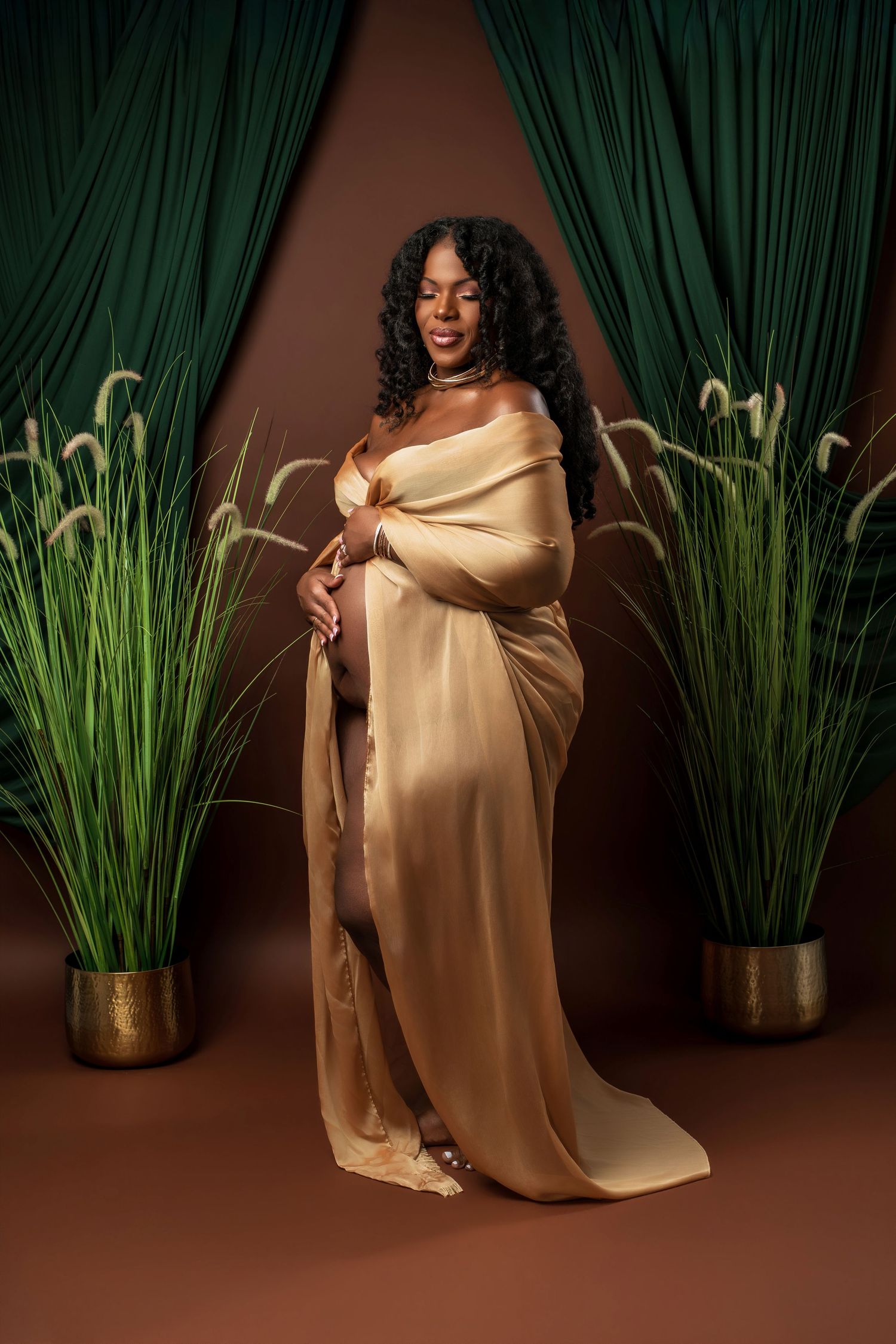A figure wrapped in a luxurious gold satin fabric poses elegantly against a dark backdrop with decorative potted plants.