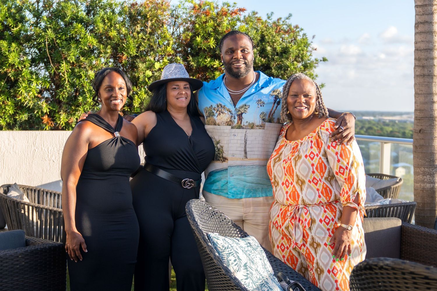 A group of people pose together on an outdoor patio with greenery and blue sky in the background.