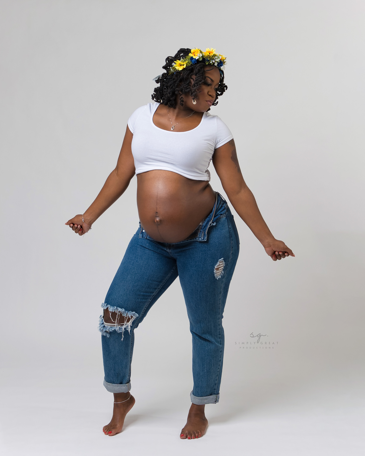 Joyful maternity pose with woman in distressed jeans and floral crown dancing.