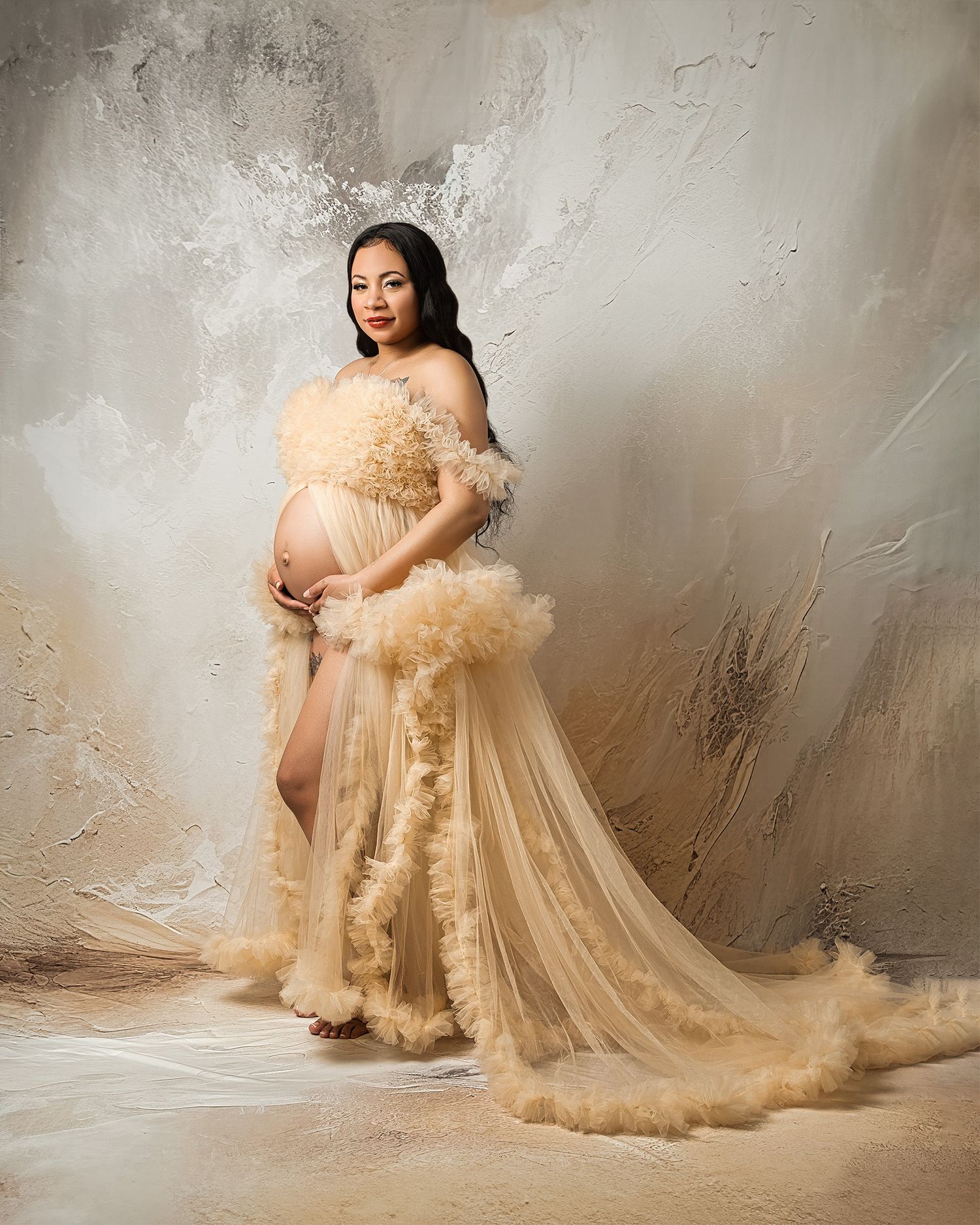Glamorous maternity portrait in champagne-colored tulle gown against textured wall.