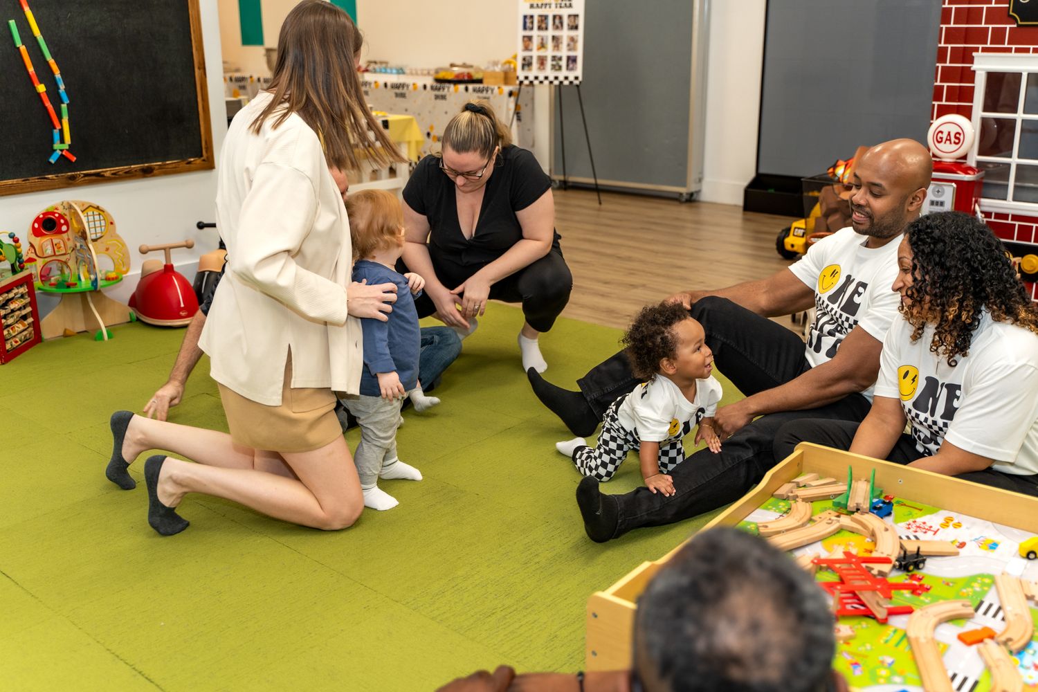 Group of adults and children gathering on green carpet for playtime activities.