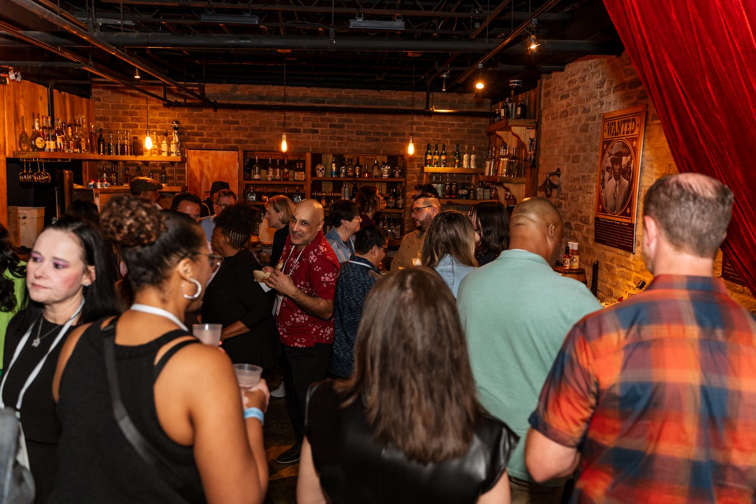 Crowded bar scene with diverse group of patrons standing near exposed brick wall.