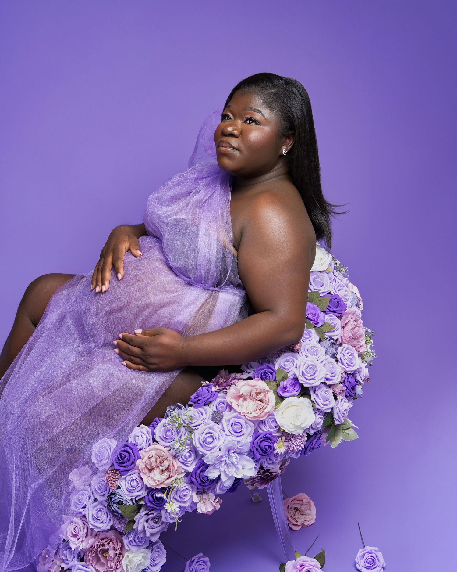 Elegant maternity pose with purple tulle dress and floral arrangement against purple backdrop.