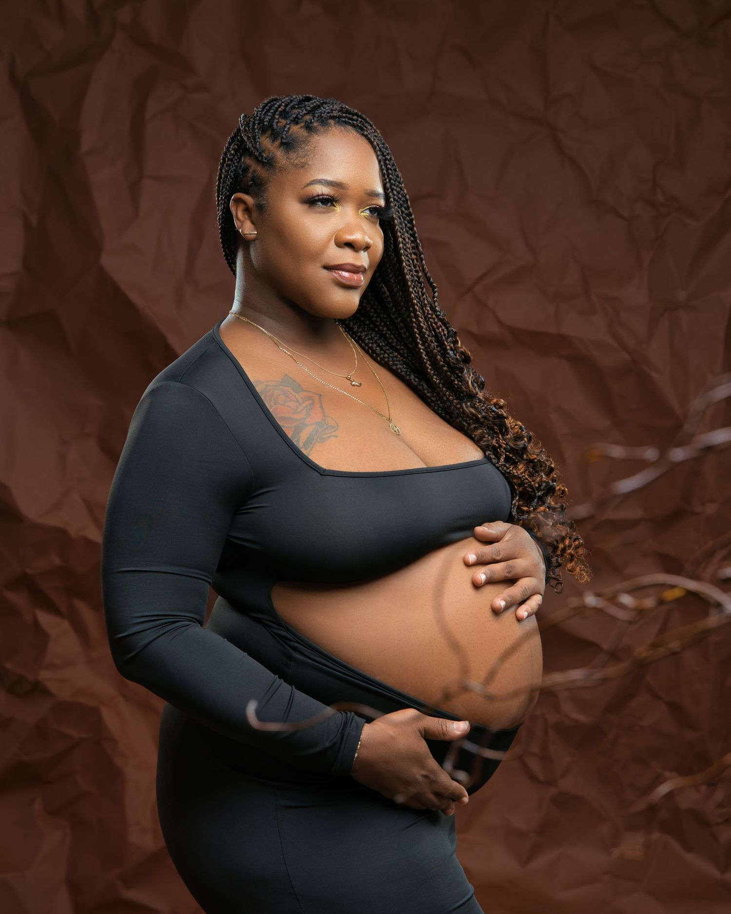 Contemporary maternity portrait against brown backdrop featuring exposed belly and long braided hair.