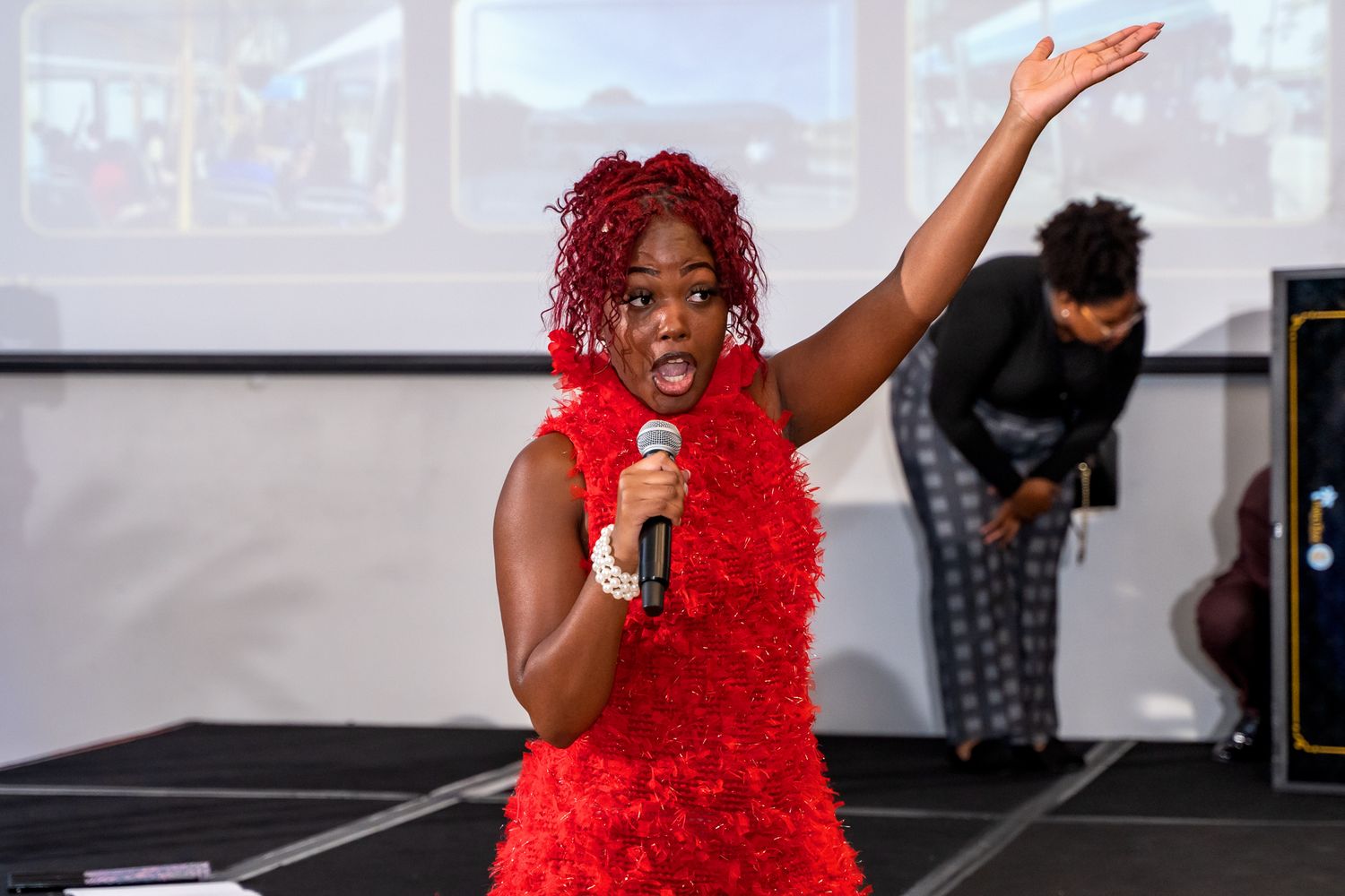 A performer in a bright red dress sings passionately on stage during an event.