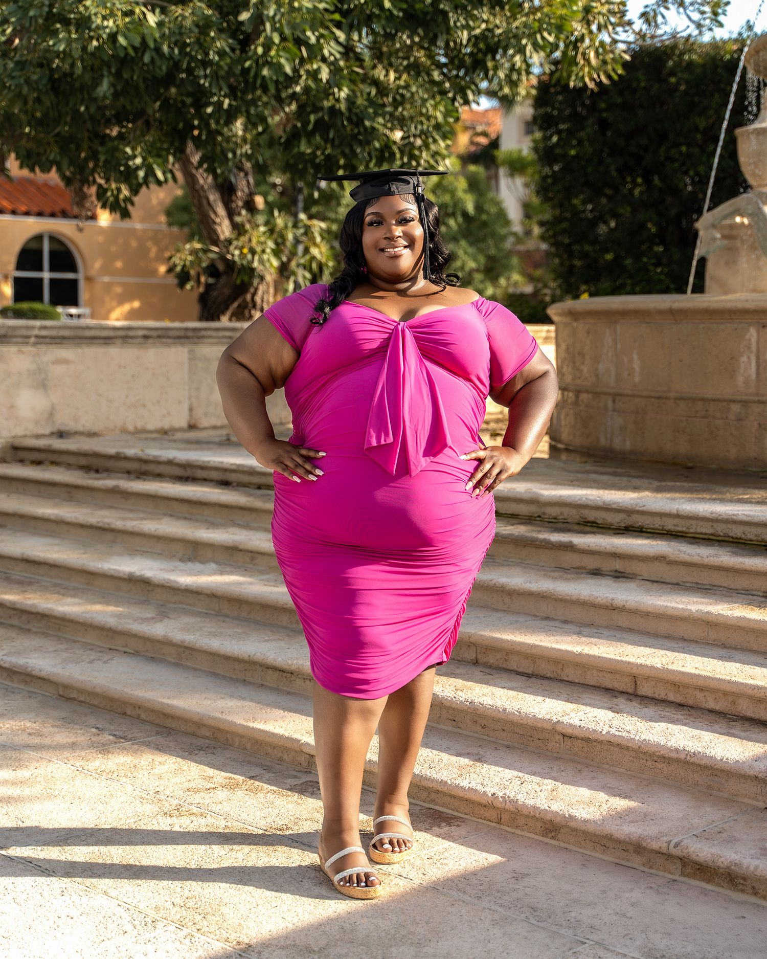 Woman in bright pink dress strikes confident pose on stone steps outdoors.