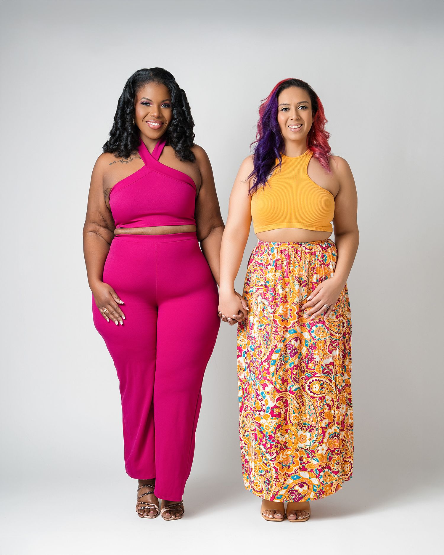 Two women pose in vibrant outfits featuring a pink jumpsuit and yellow top with patterned skirt.