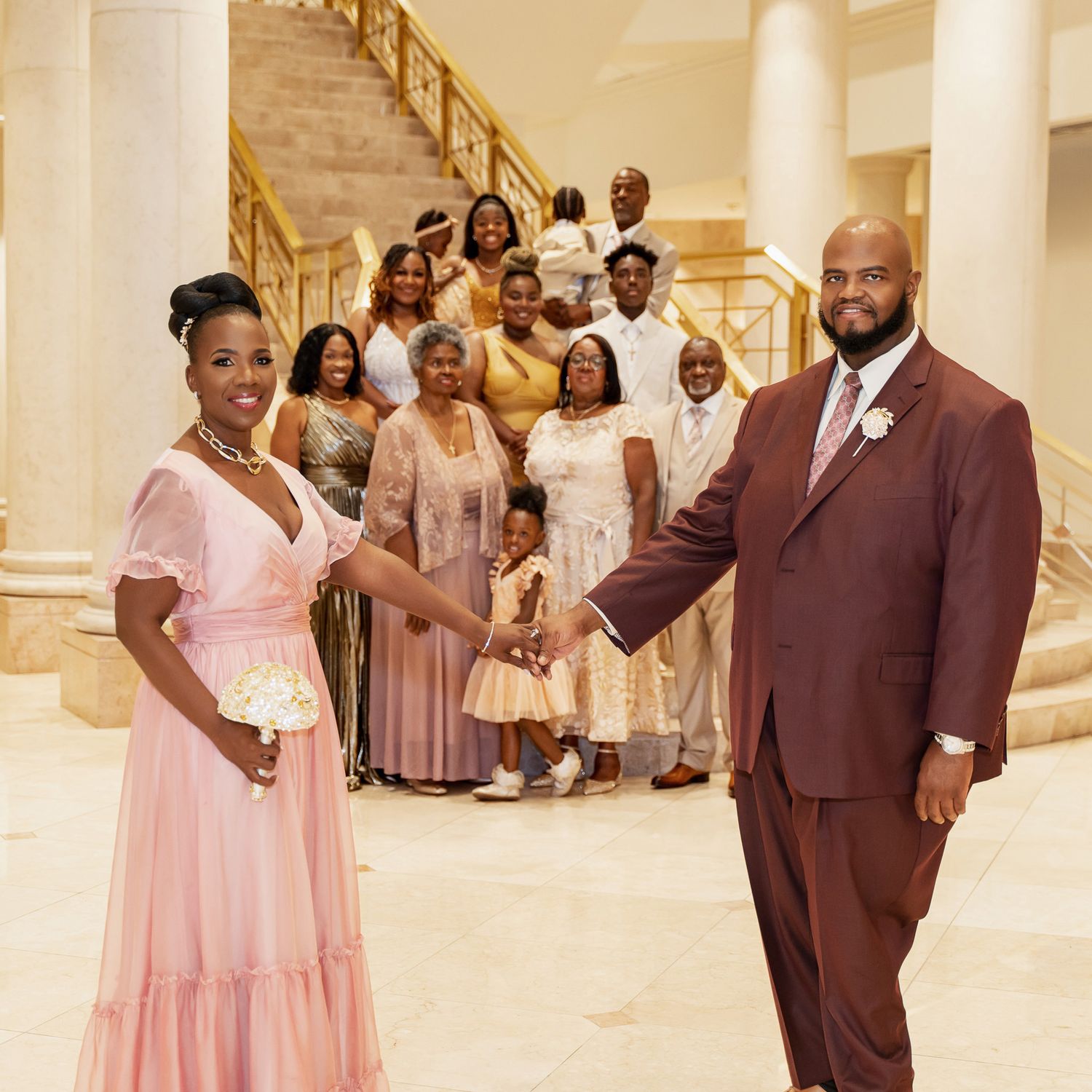 A wedding party posing on grand marble stairs in formal attire with pink and brown color scheme.