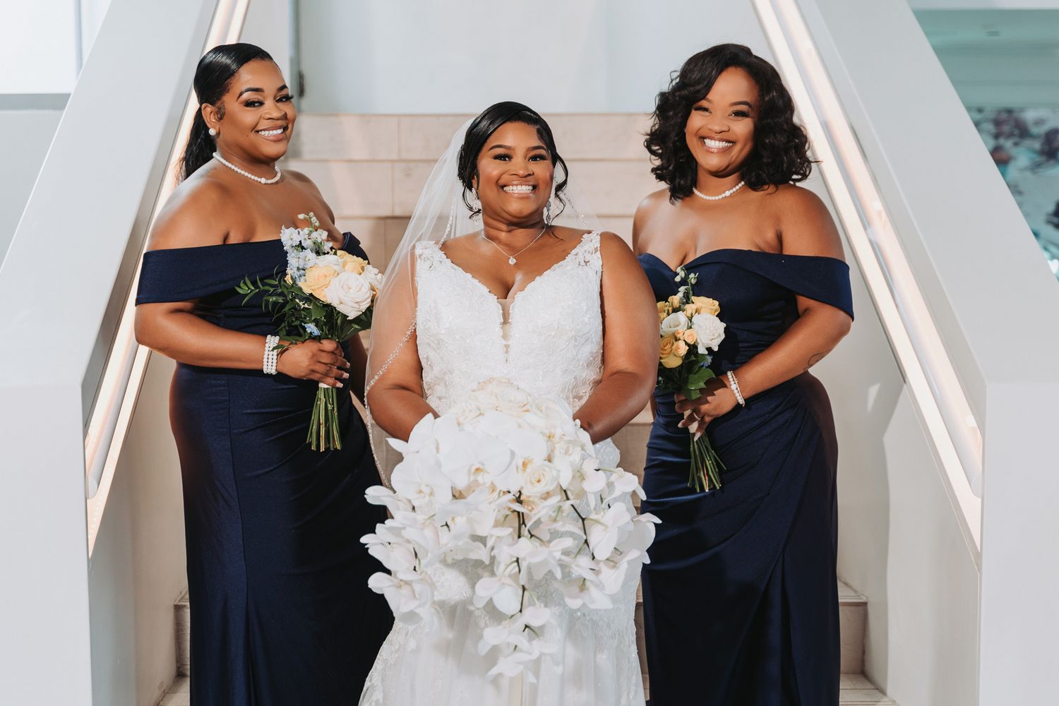 Three women in elegant navy blue gowns pose together with white floral bouquets.