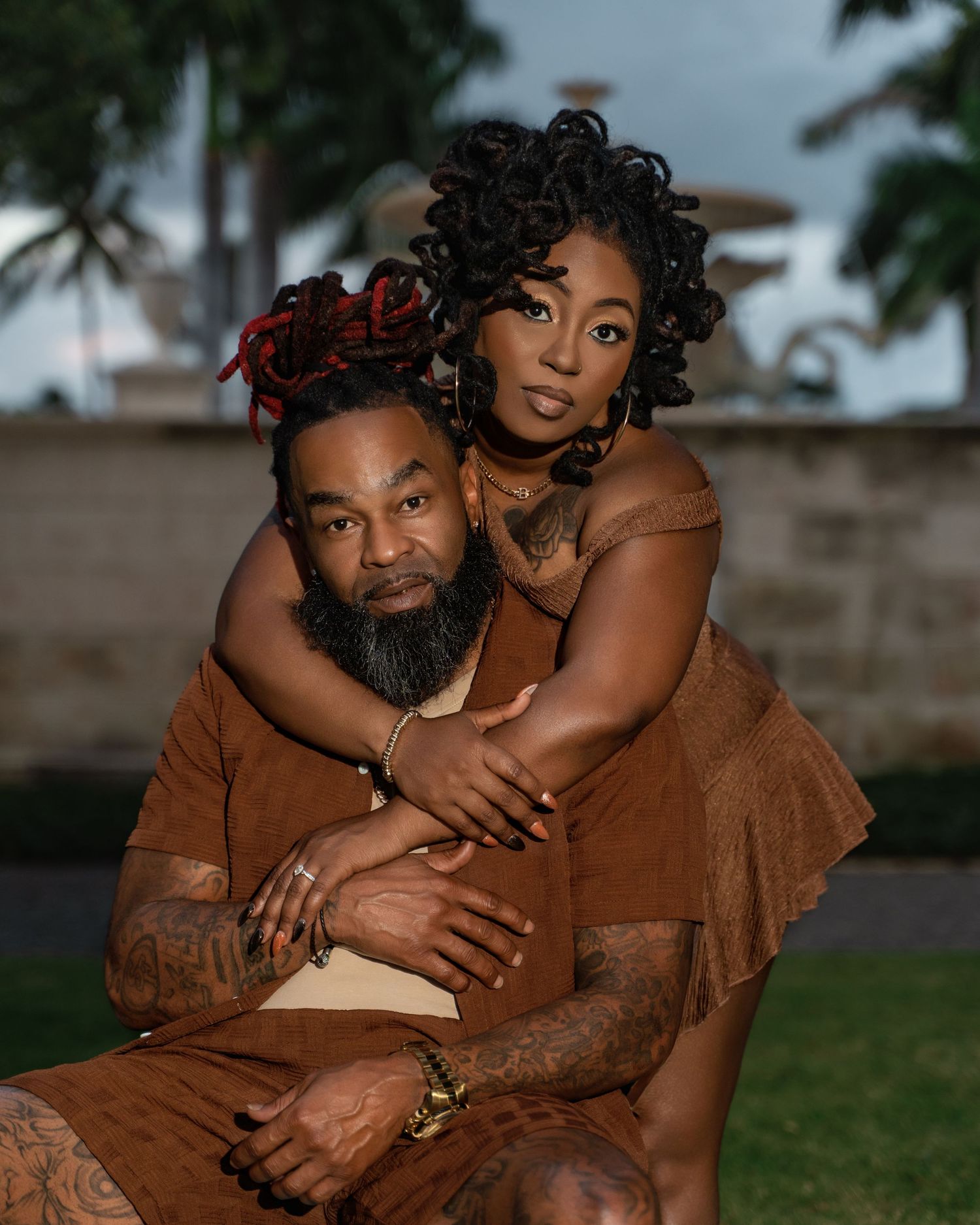 A romantic outdoor portrait of a couple embracing in brown attire with palm trees in the background.
