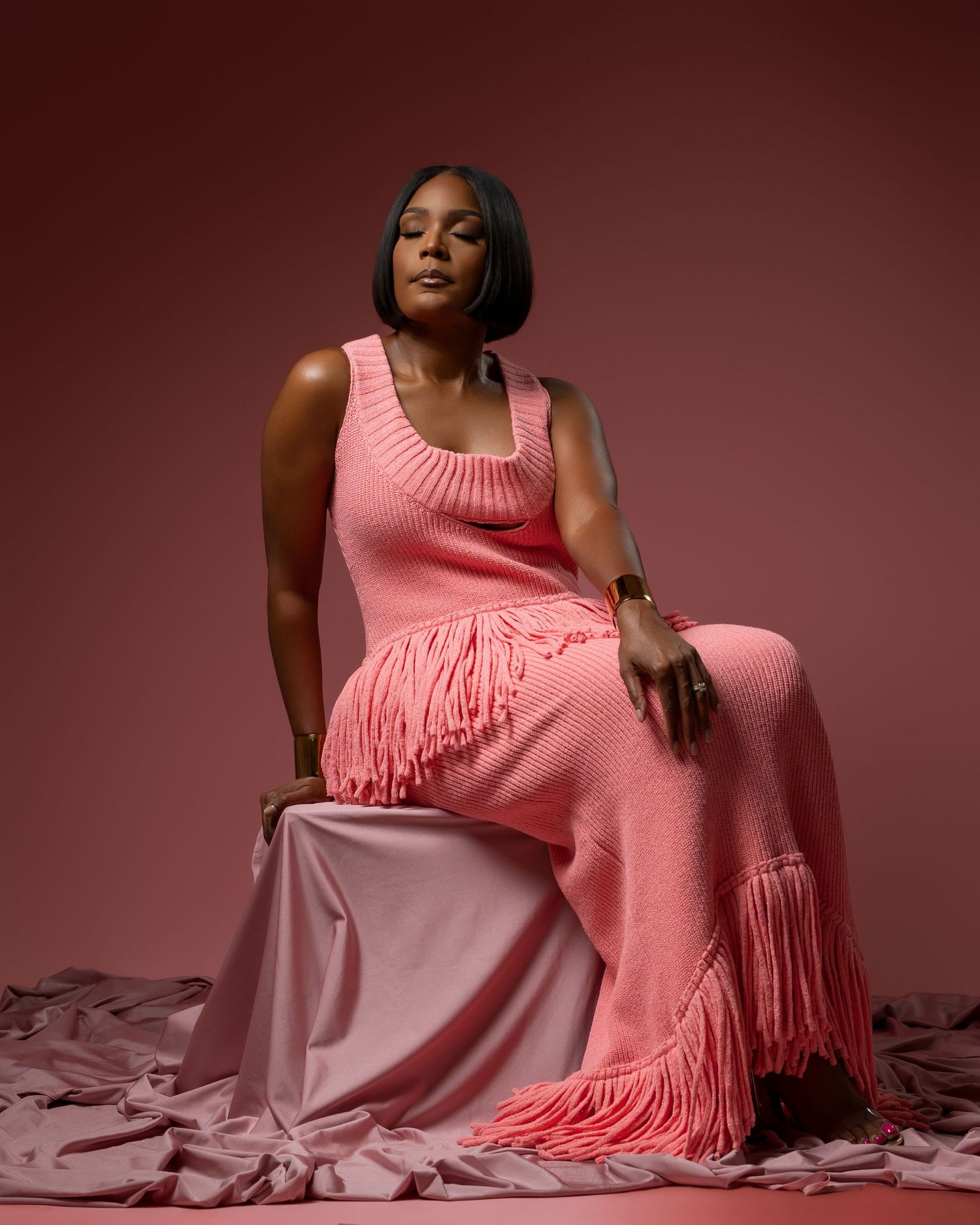 A figure in a pink sweater dress with fringe detail poses gracefully against a burgundy backdrop in a photo series.