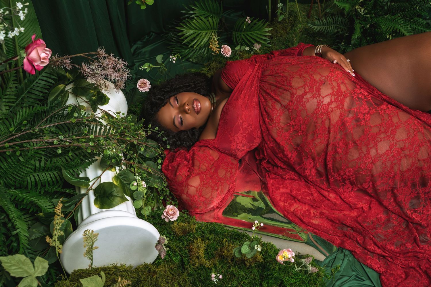 A person in red lace lounges among lush green foliage and pink flowers in a garden setting.