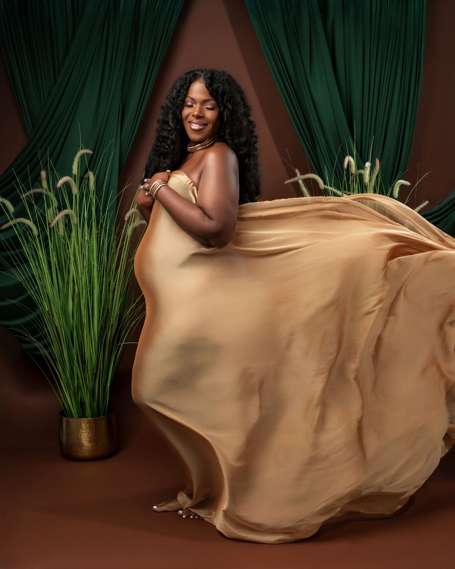 A flowing gold silk fabric creates elegant movement against a dark backdrop with green plants.