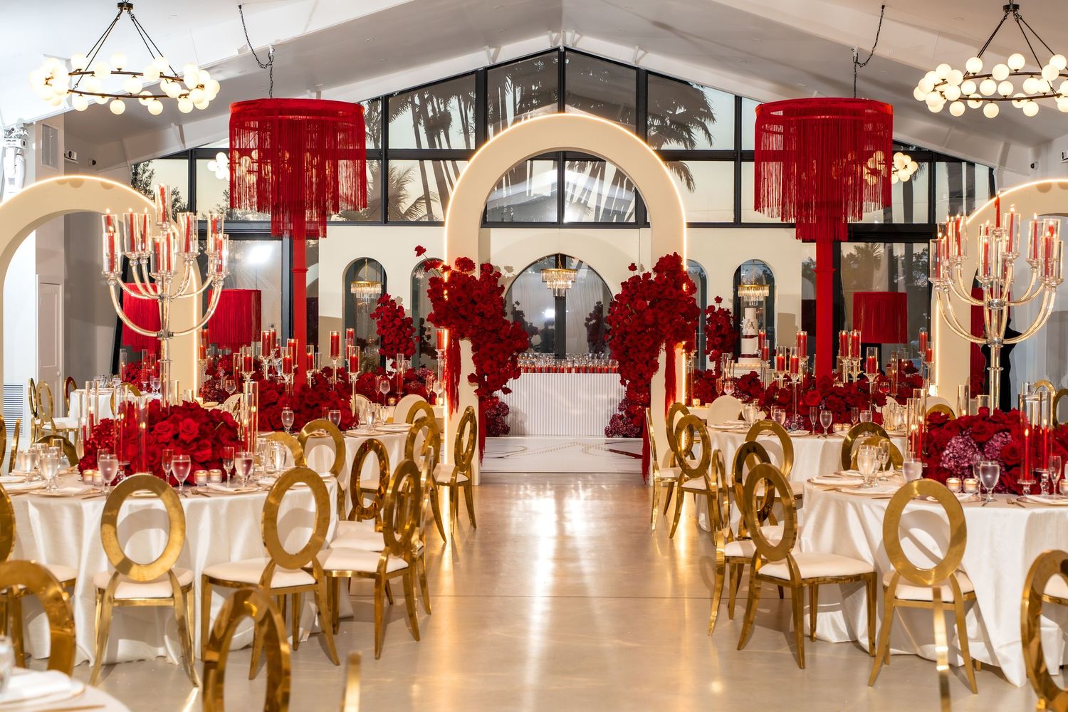 An elegant wedding reception venue with gold chairs and red floral centerpieces arranged in a long banquet hall setup.