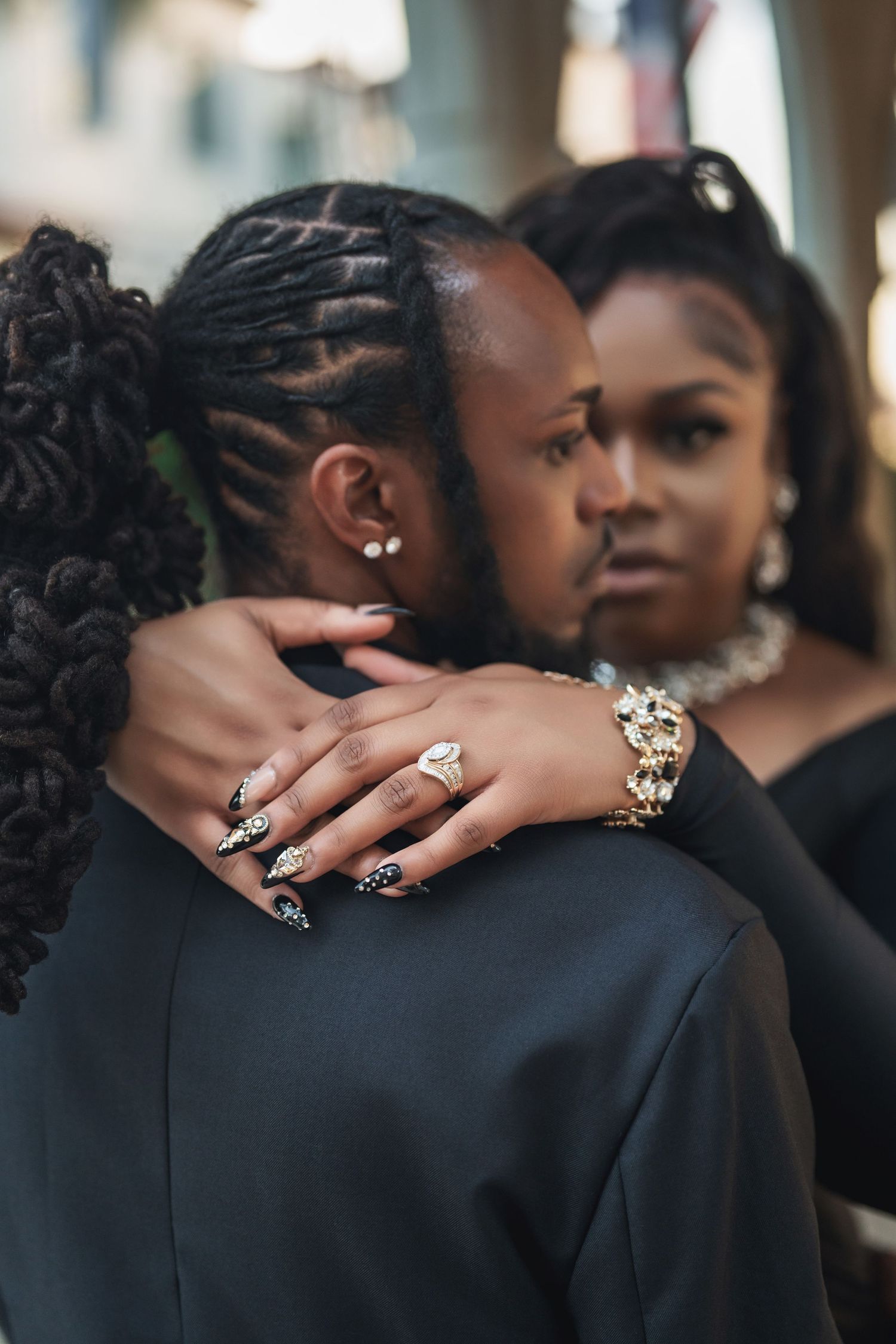 Two people share an intimate embrace in formal black attire, with one person displaying elegant jewelry and nail art.