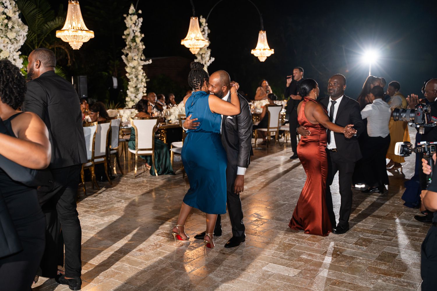 Outdoor evening celebration with couples dancing under illuminated chandeliers.