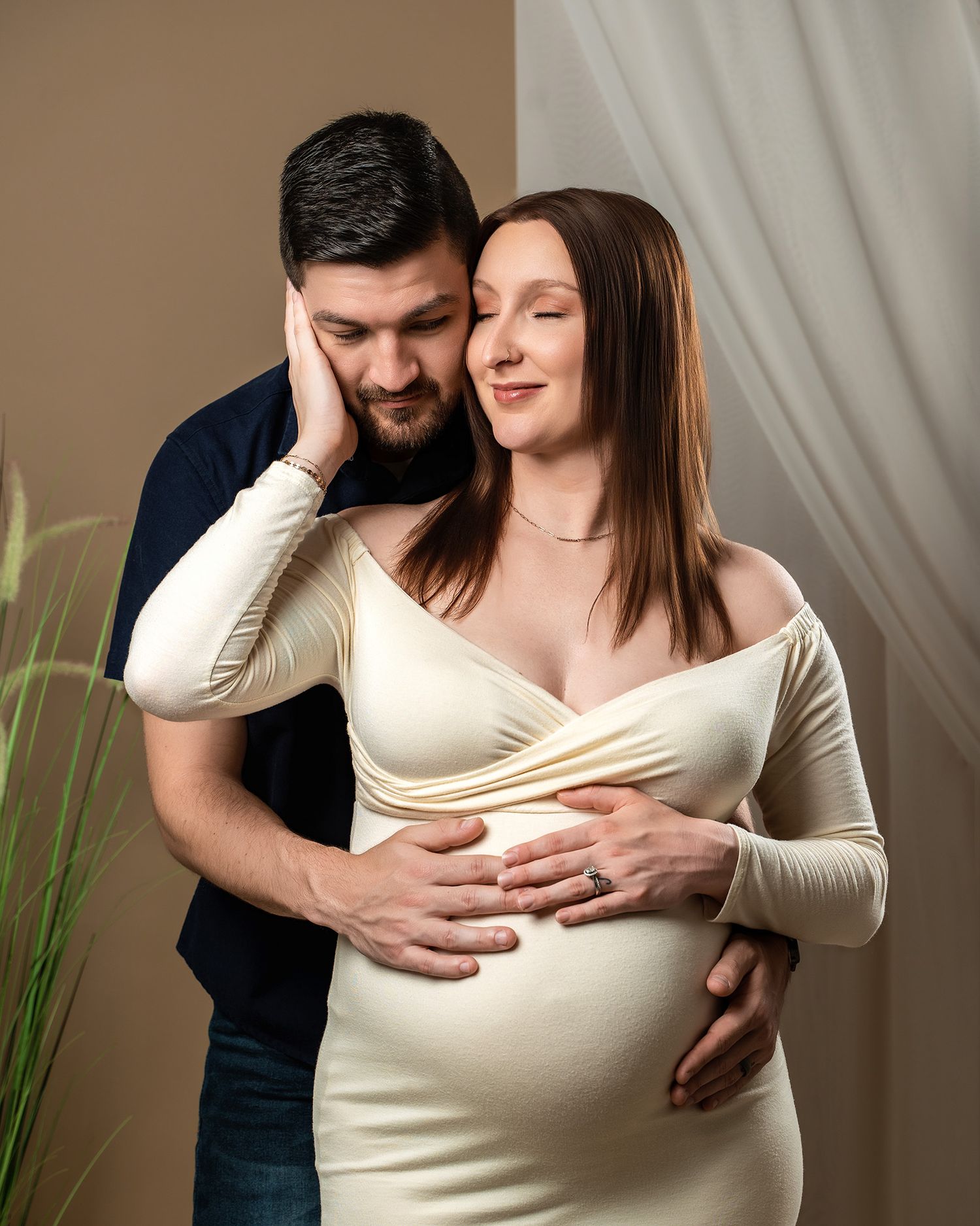 Tender maternity portrait with couple embracing near decorative plants and cream curtains.