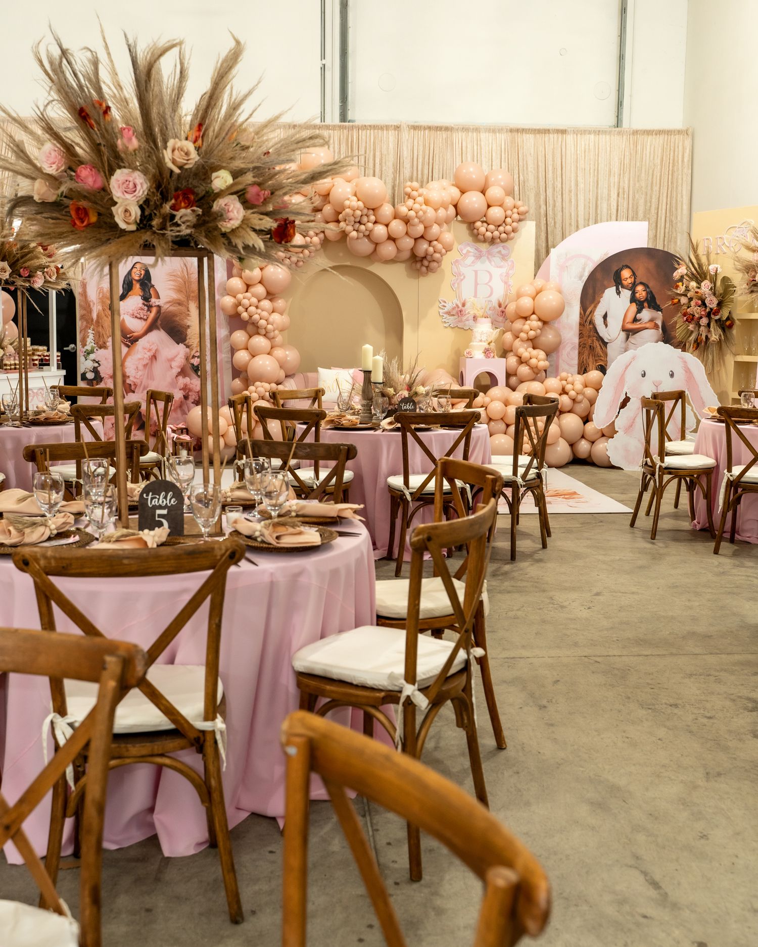 Elegant baby shower venue with pink table settings, balloon decor, and rustic wooden chairs.
