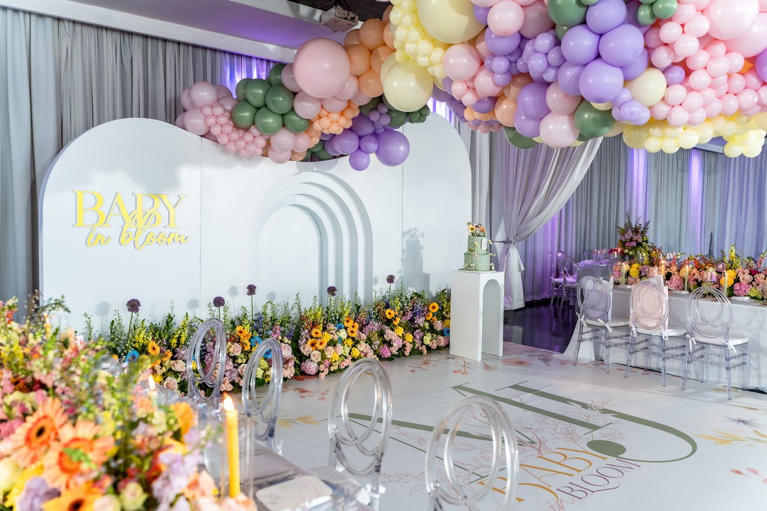 Modern baby shower venue with pastel balloon garland, white arch backdrop and floral arrangements.