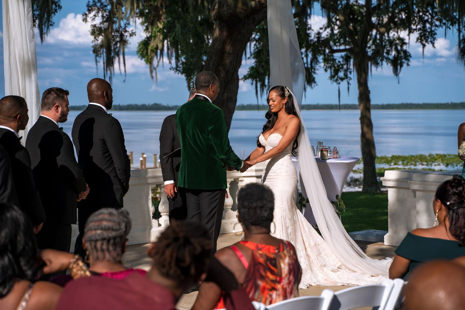 An outdoor waterfront wedding ceremony with guests seated on white chairs.