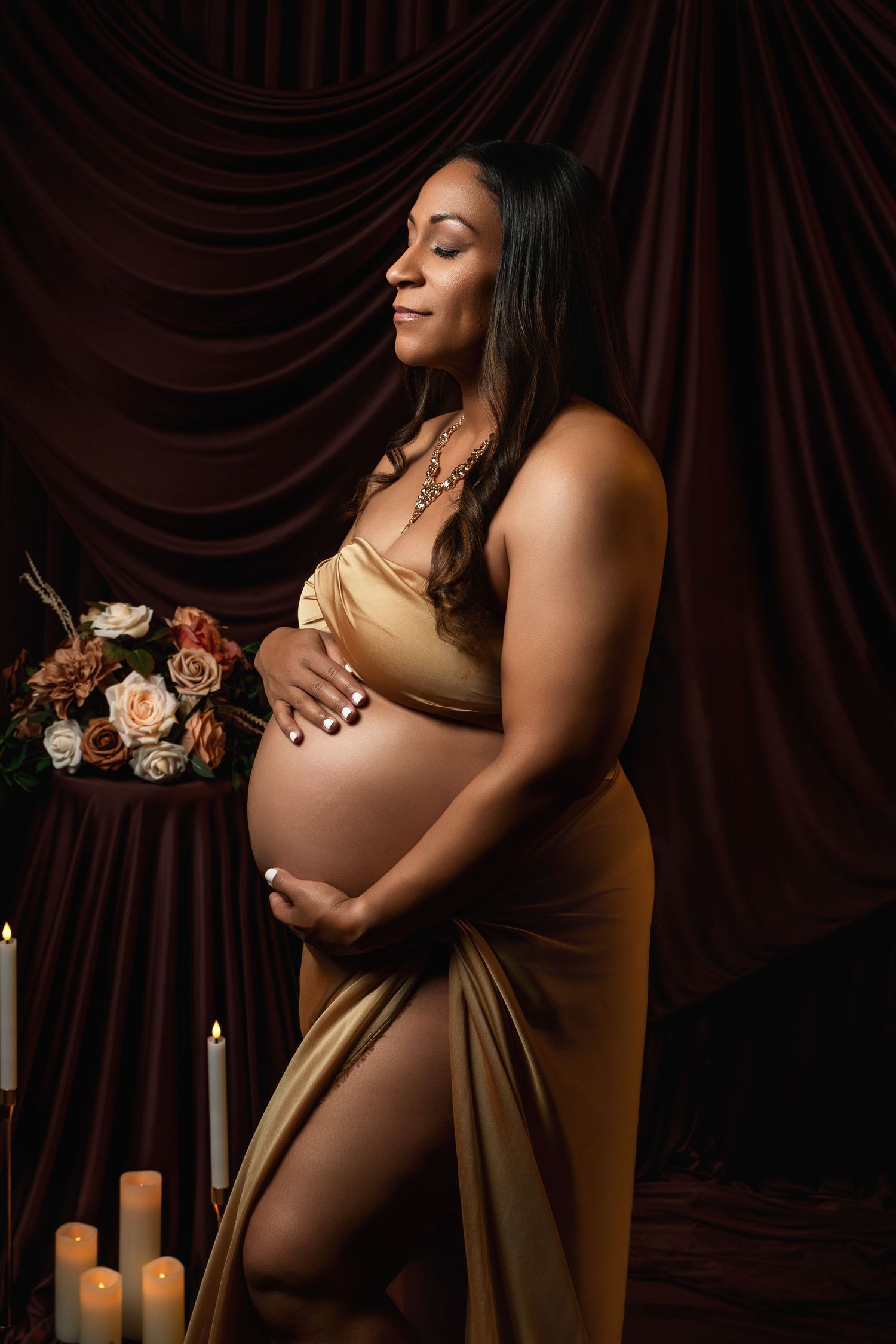 Maternity photoshoot with dramatic brown drapes, candles, and floral arrangements creating an intimate atmosphere.