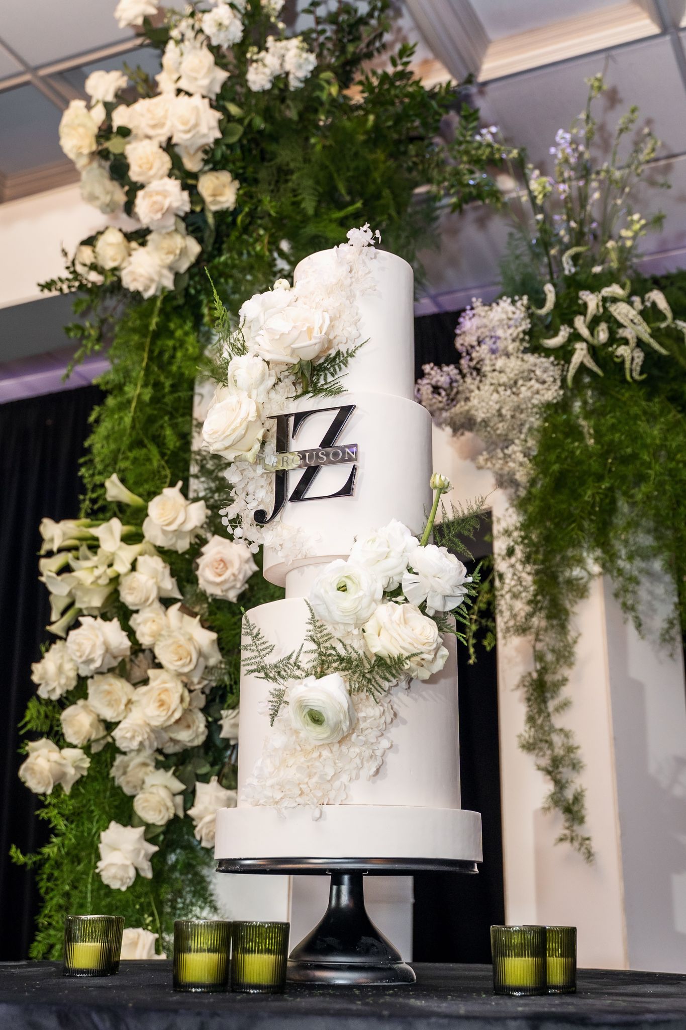 Three-tiered white wedding cake displayed with lush floral arrangements and green votives.