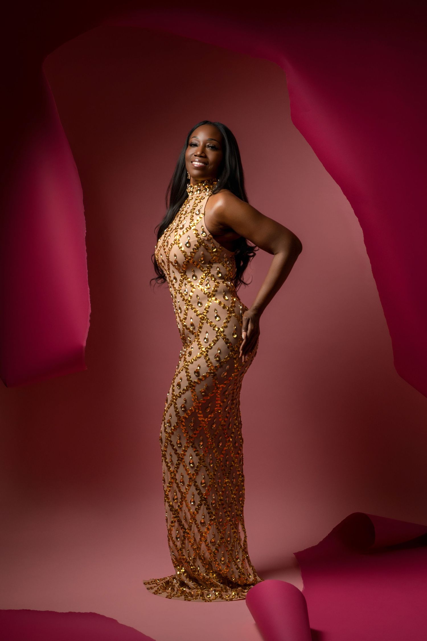 A person poses confidently in a floor-length gold sequined gown against a deep burgundy background.