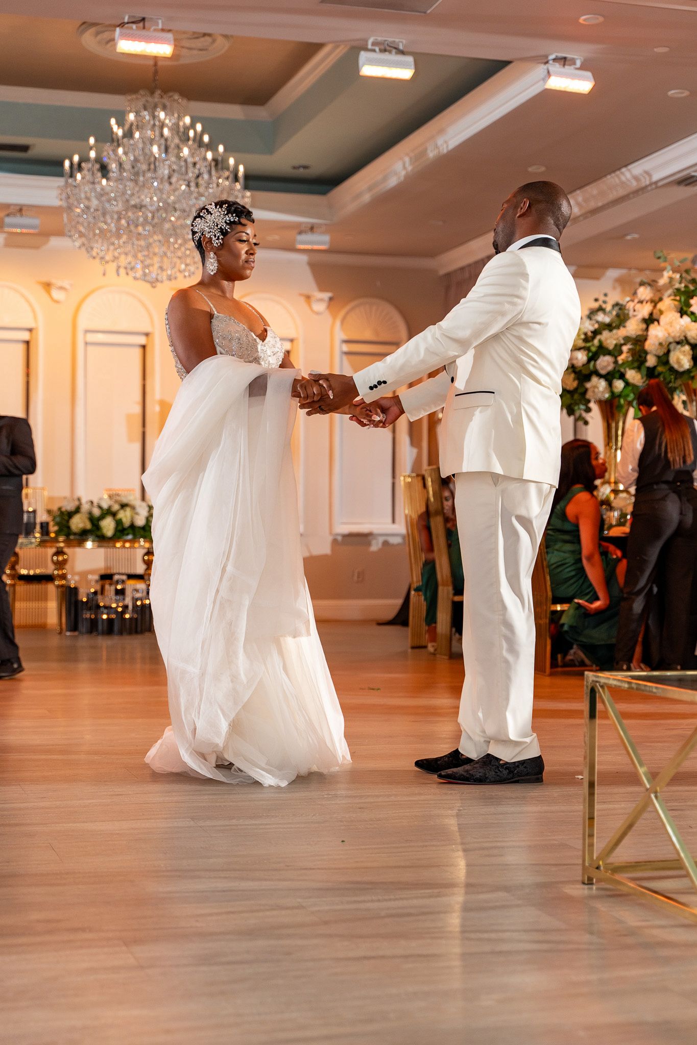 Couple dances gracefully in white attire on a hardwood floor beneath crystal chandeliers.