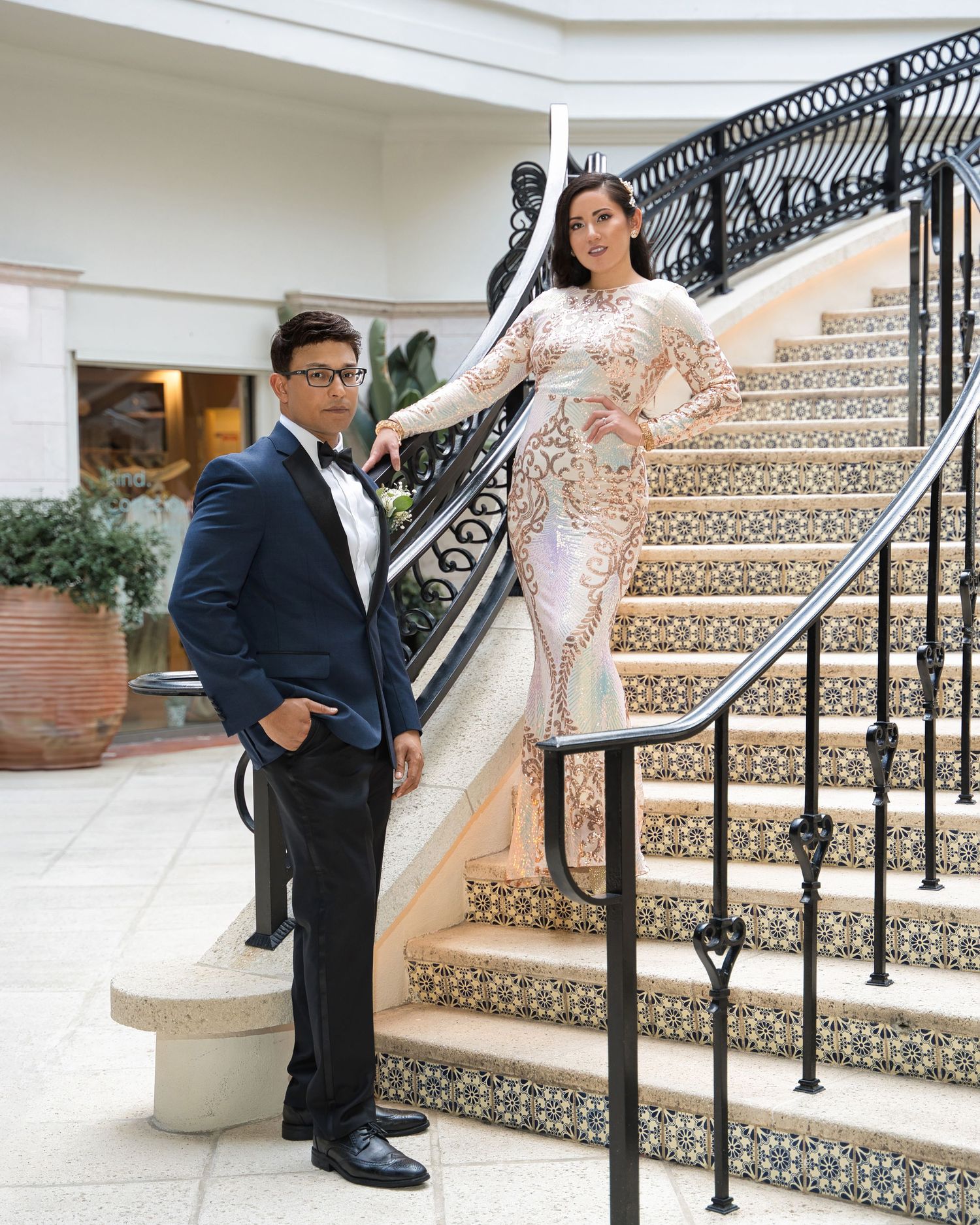 Elegant couple poses on ornate staircase with decorative railing, bride wearing fitted lace gown.