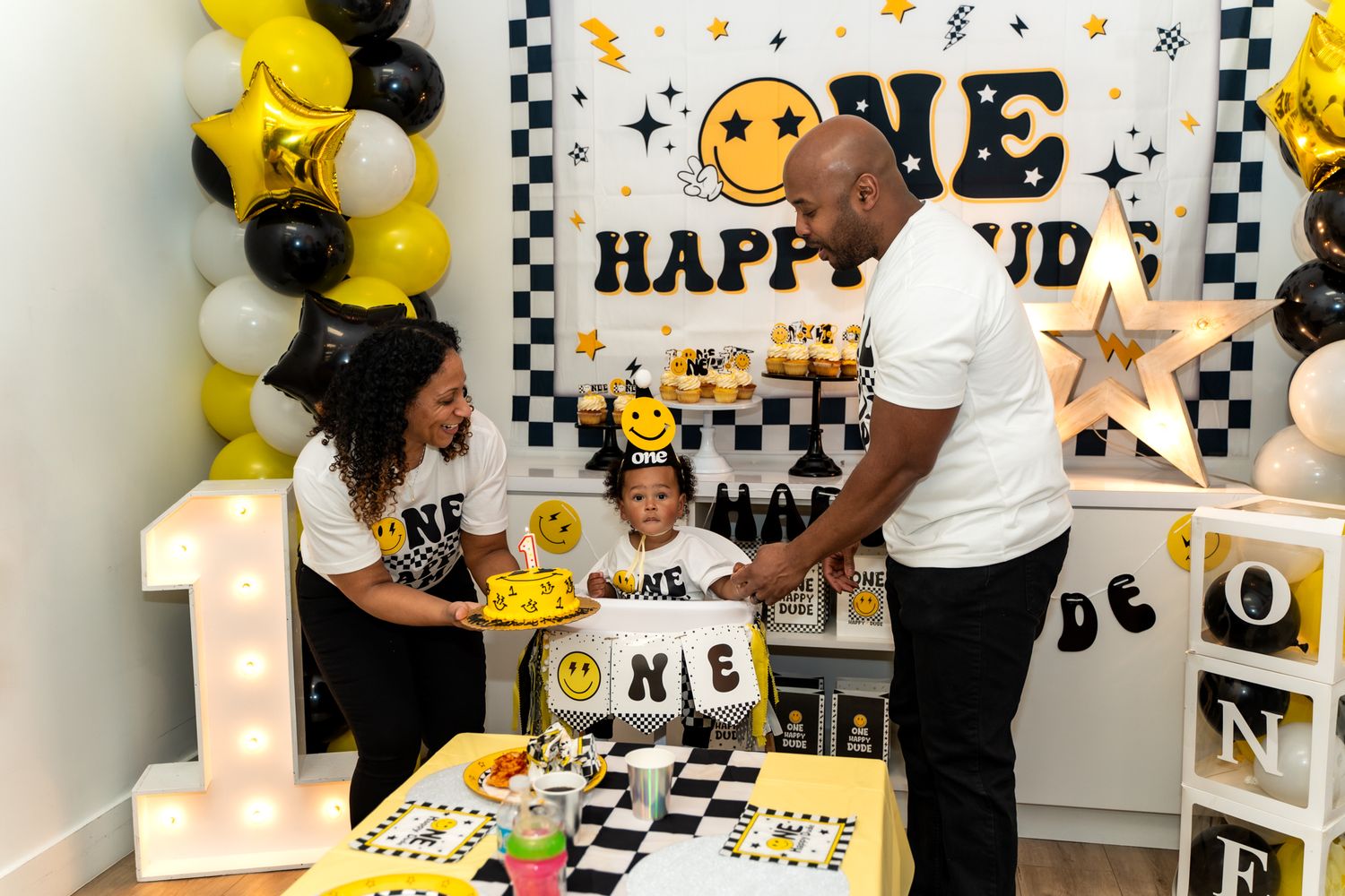 A festive first birthday party setup with yellow and black balloons and a smiley face theme featuring a large number 1 decoration.