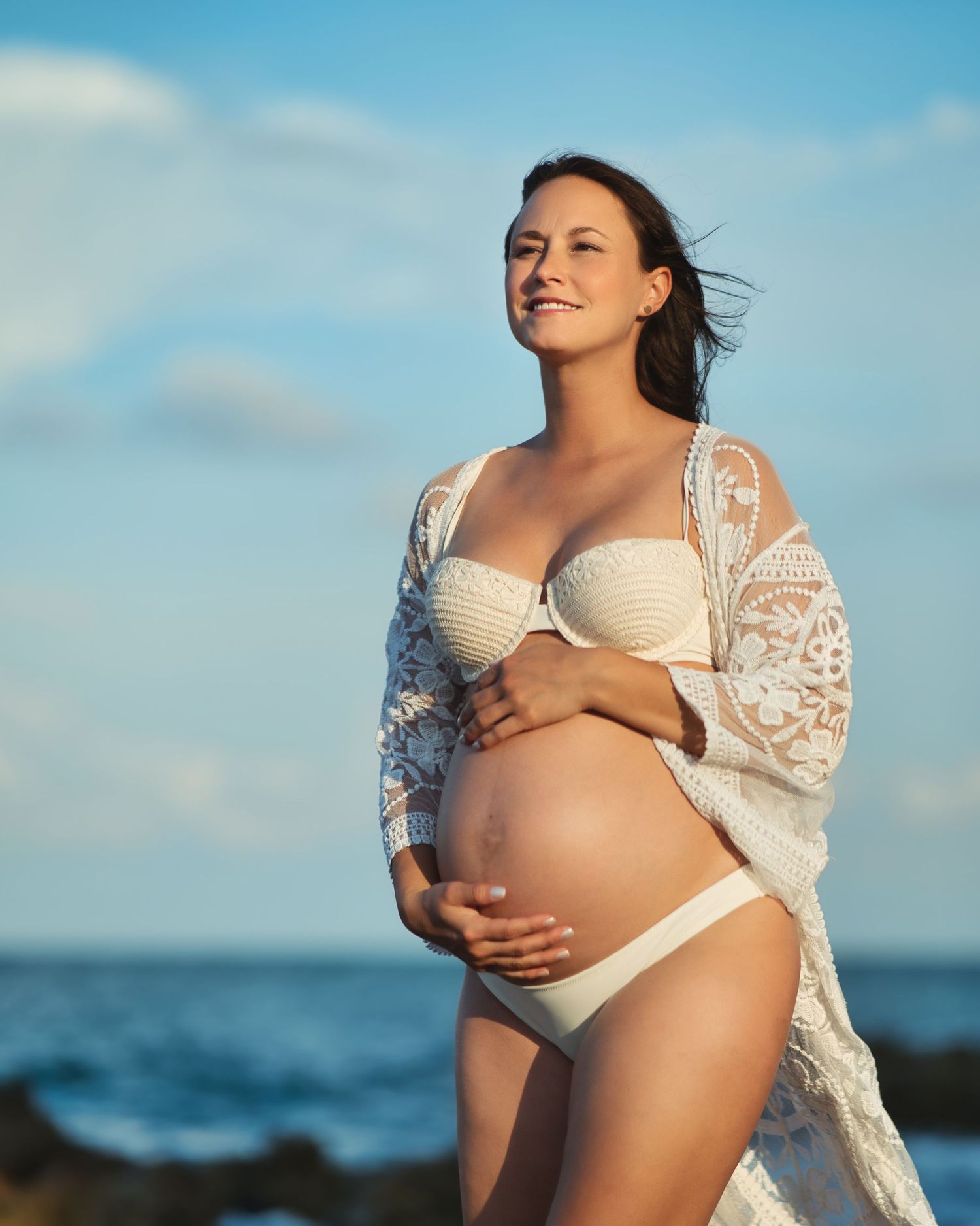 Maternity beach photoshoot featuring expecting mother in white swimwear and lace coverup against ocean backdrop.