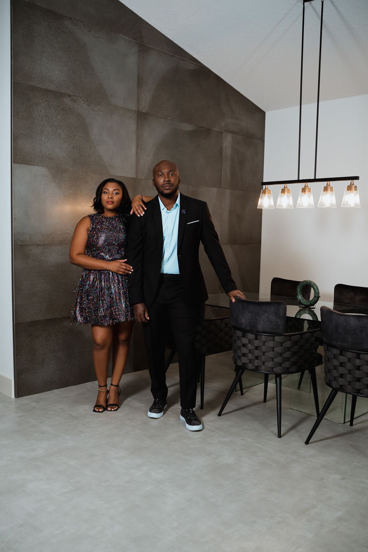 Elegantly dressed couple standing against textured gray wall in modern interior setting.