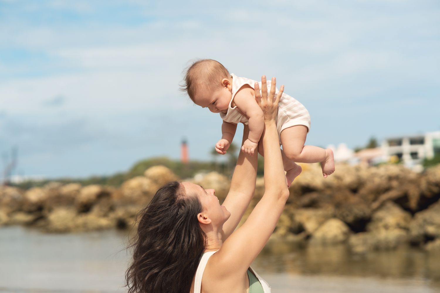 A parent lifts a smiling baby high into the air at a scenic beach with rocks and buildings in background.