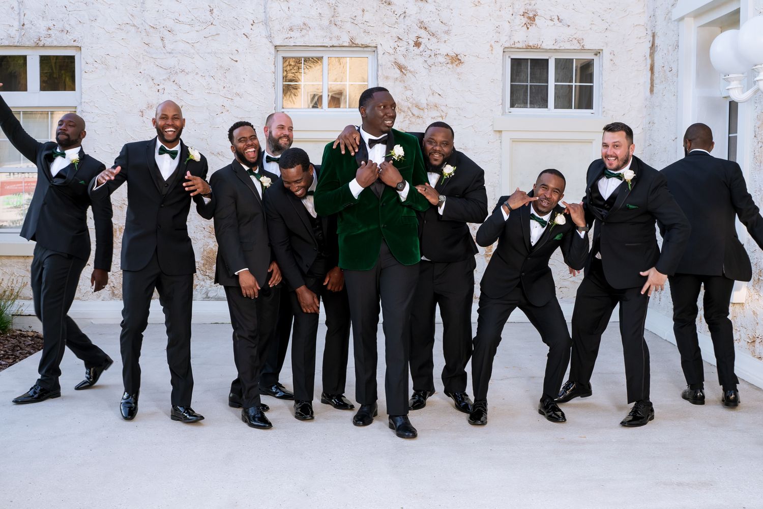 Groomsmen in black suits and one in green velvet celebrate with playful poses against a white wall.
