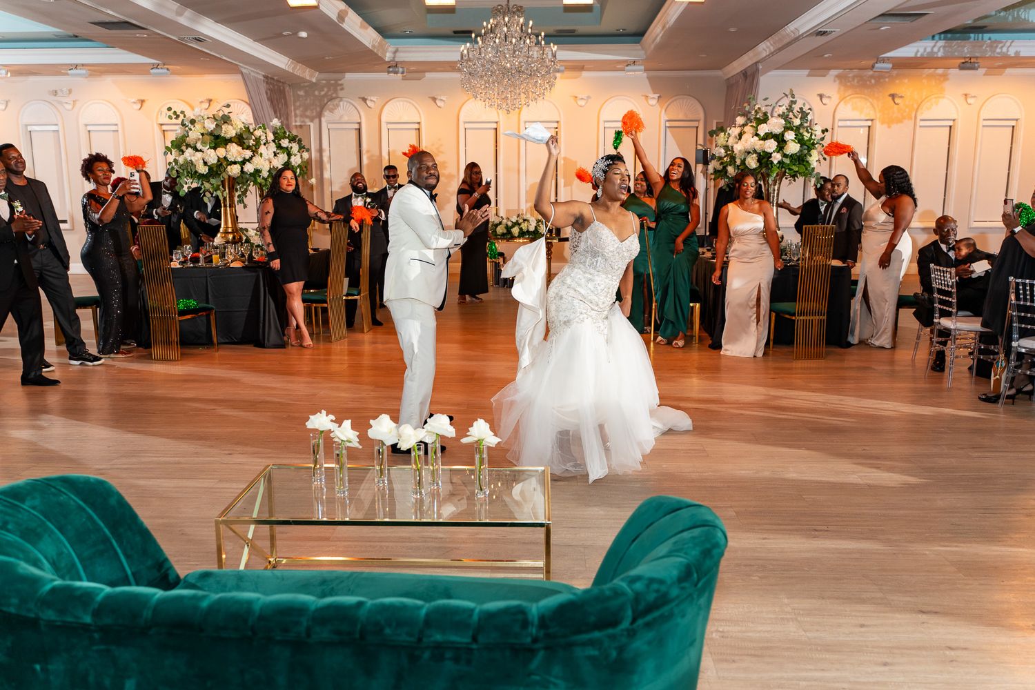 Newlyweds share their first dance beside a green velvet sofa in an elegant ballroom with floral decor.