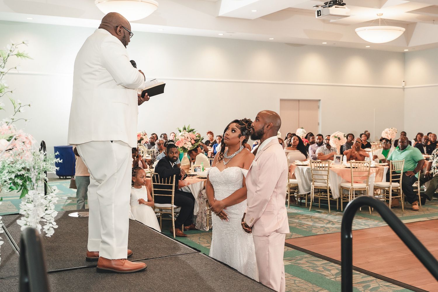 Wedding ceremony in progress with guests seated in a reception hall with pink and white decor.