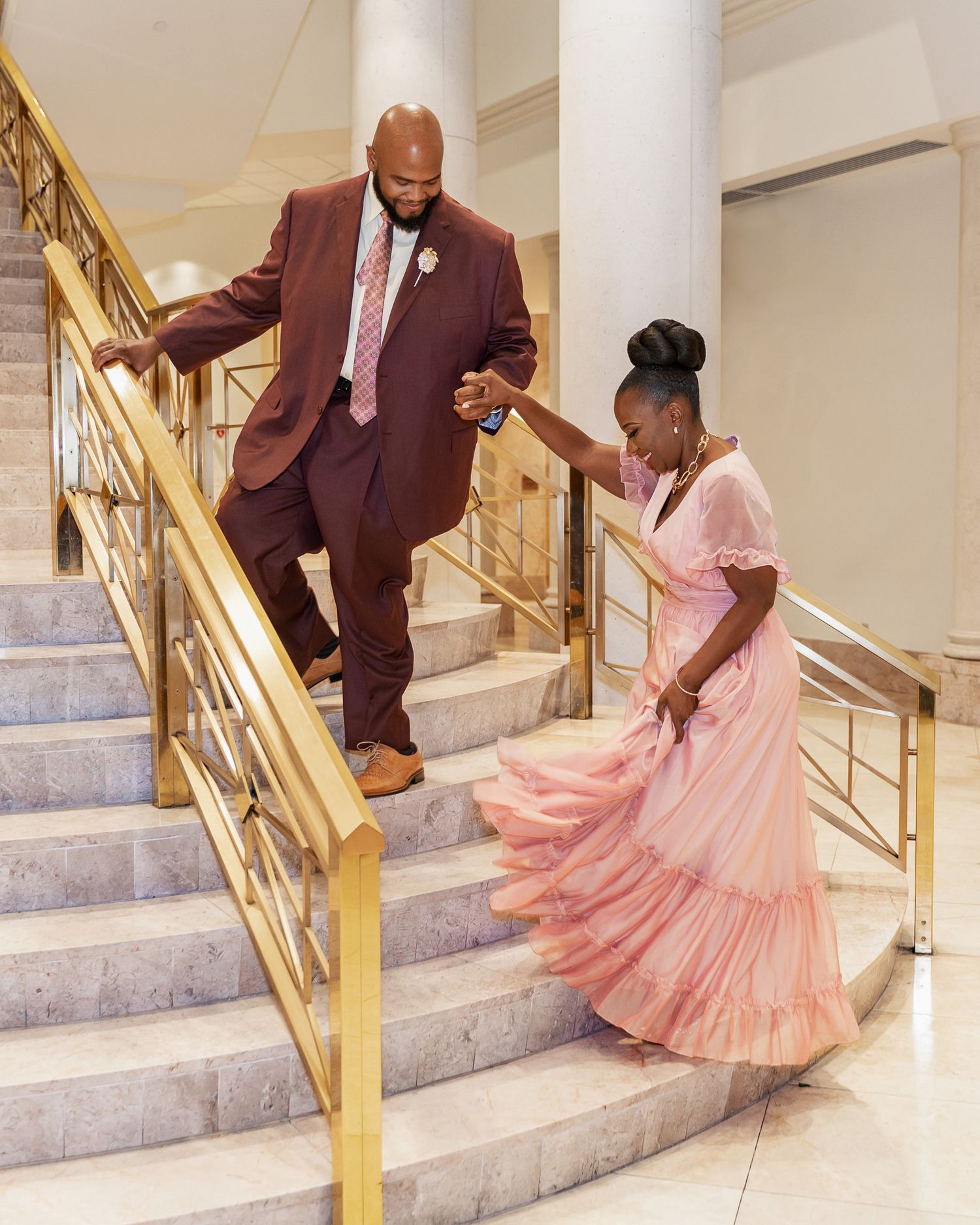 A couple descends marble stairs together, her pink dress flowing as they hold hands.