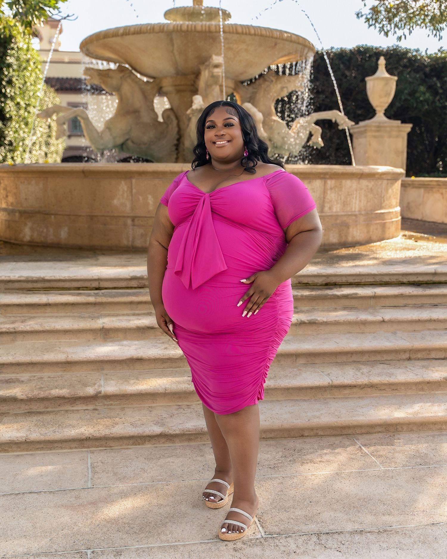 Woman wearing a pink dress with bow detail stands on steps near a flowing fountain.