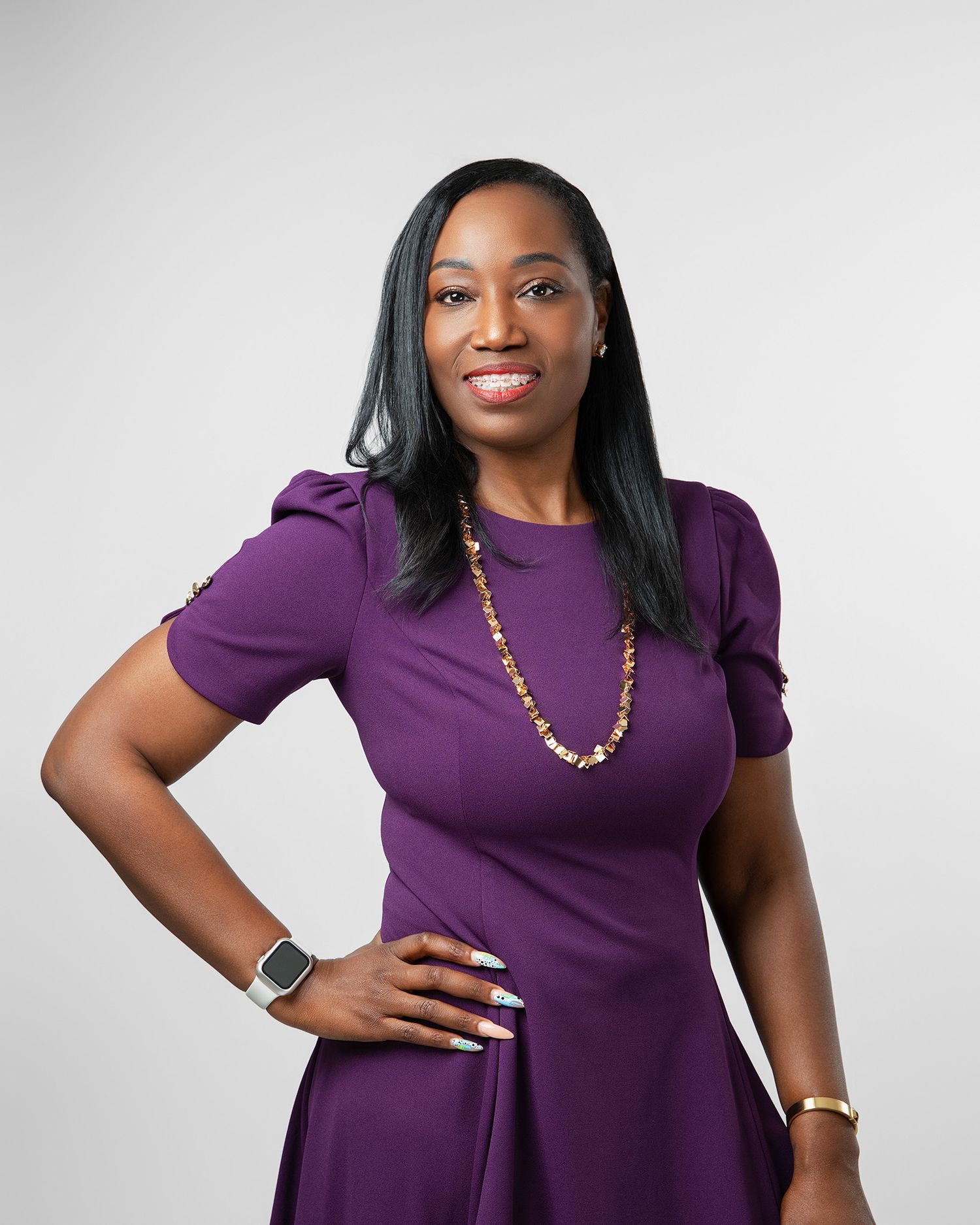 Professional headshot of someone wearing a purple dress and beaded necklace against a light gray background.