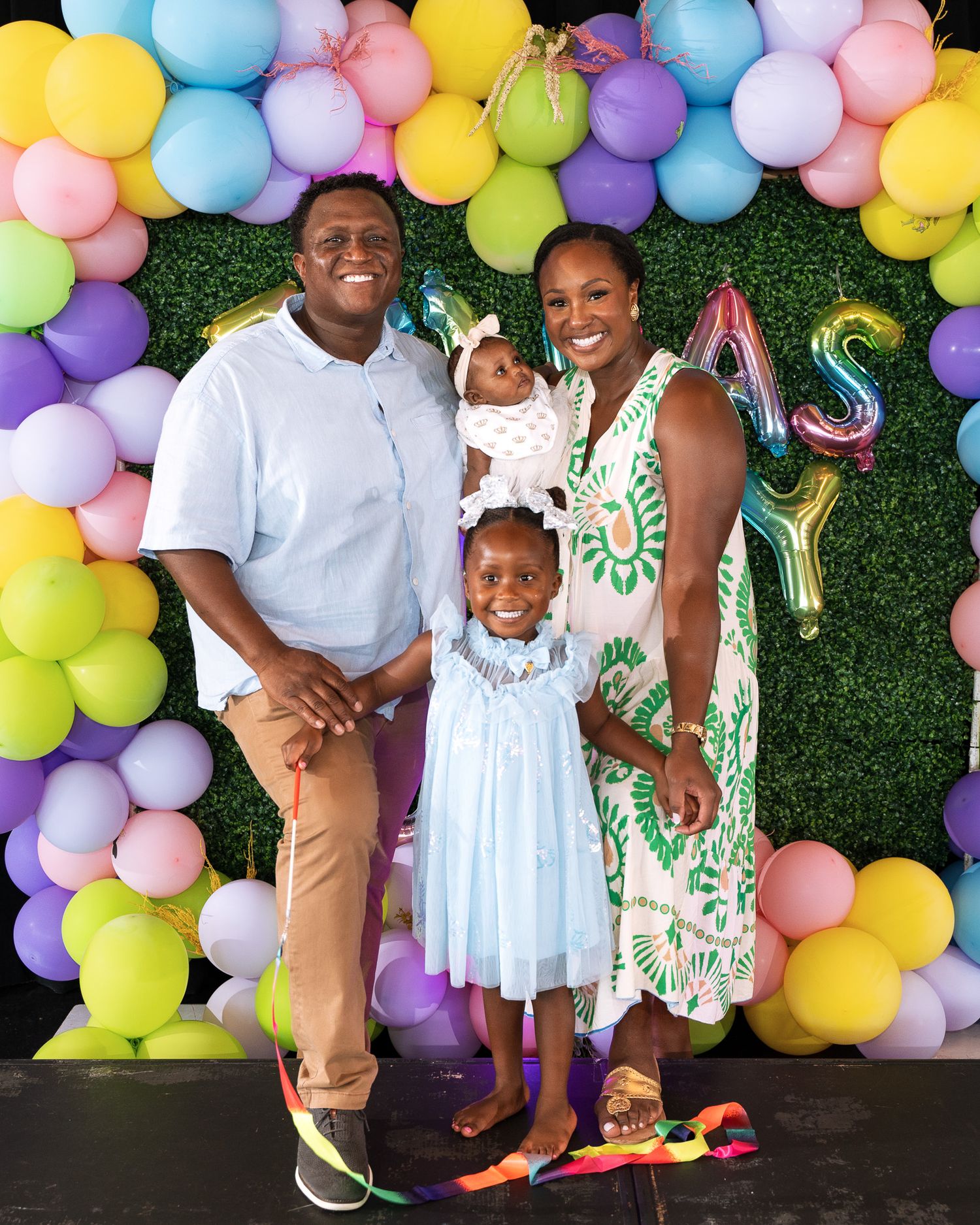 A family portrait against a backdrop of colorful balloons and greenery at a celebration.
