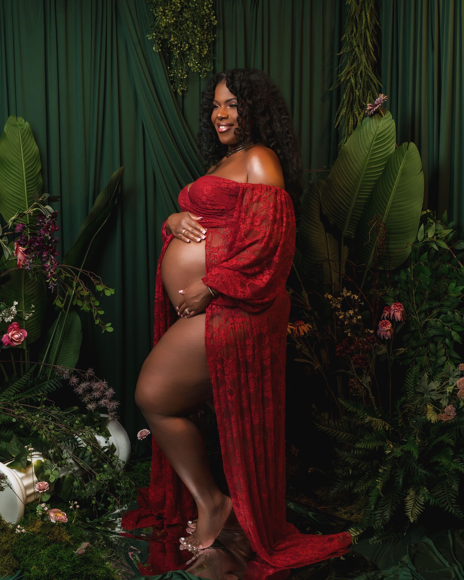A maternity photoshoot features a flowing red dress against a lush green botanical backdrop with dark curtains.